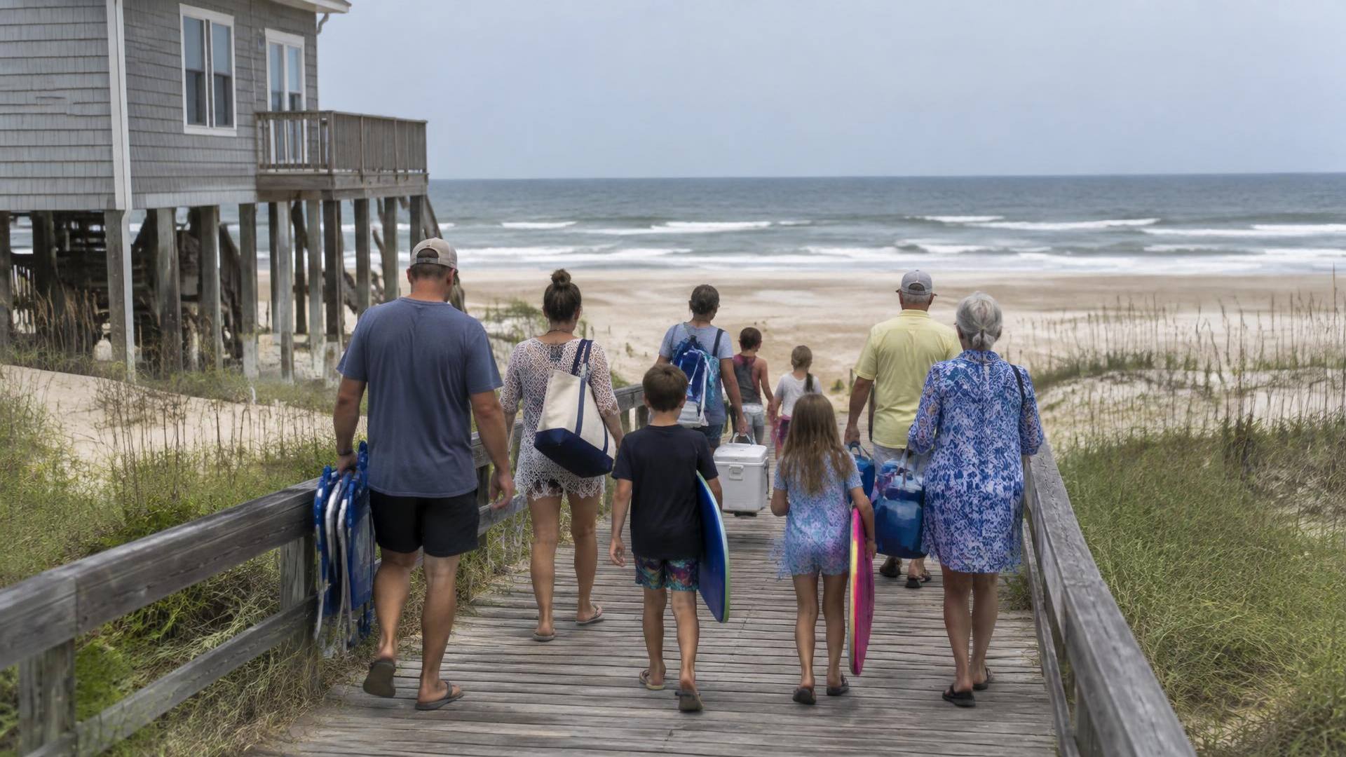 Multi-generational family carrying chairs, a cooler and boogie boards down a beach walkway