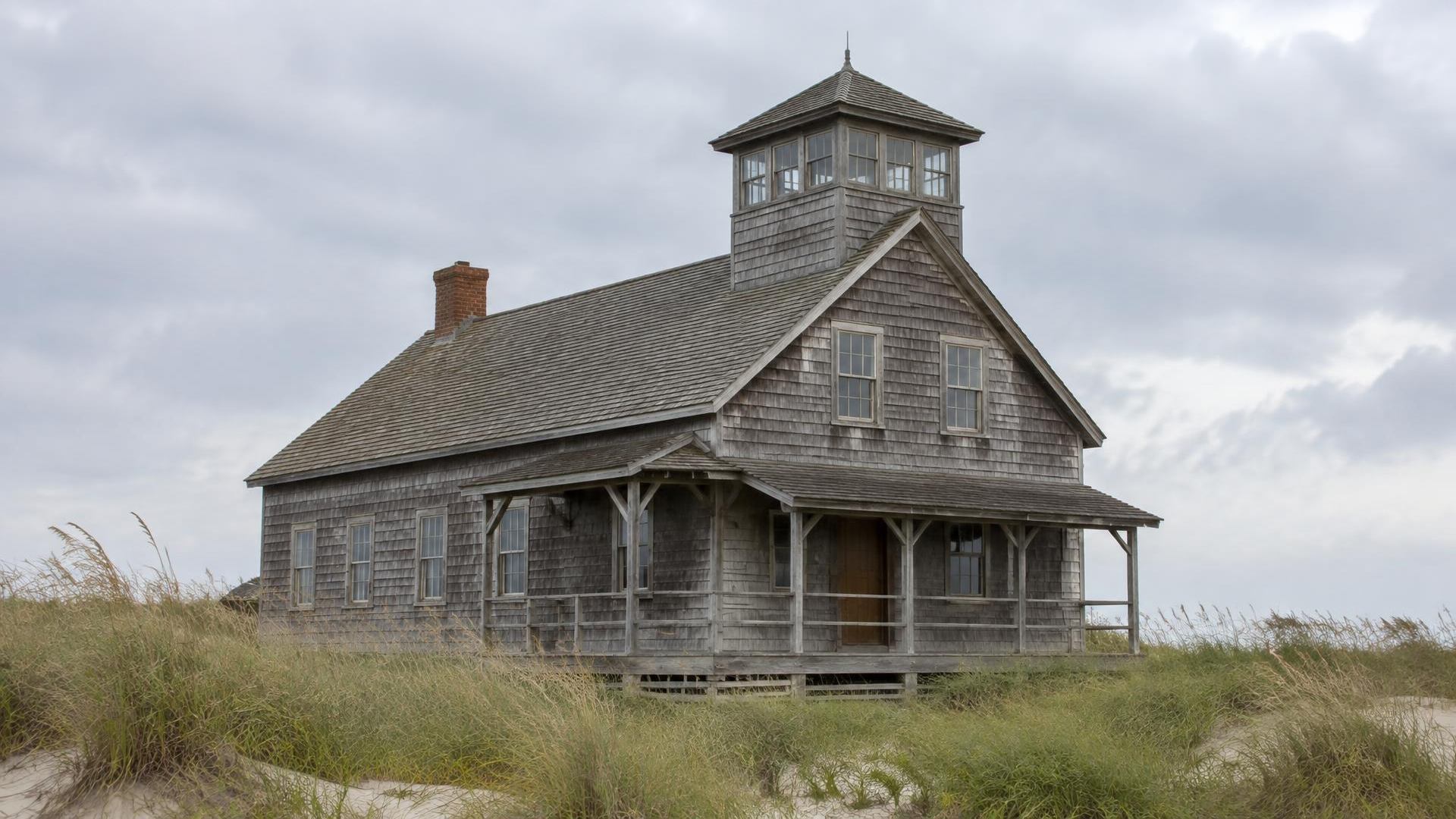 Weathered wooden life-saving station with a lookout tower among dune grass under overcast skies